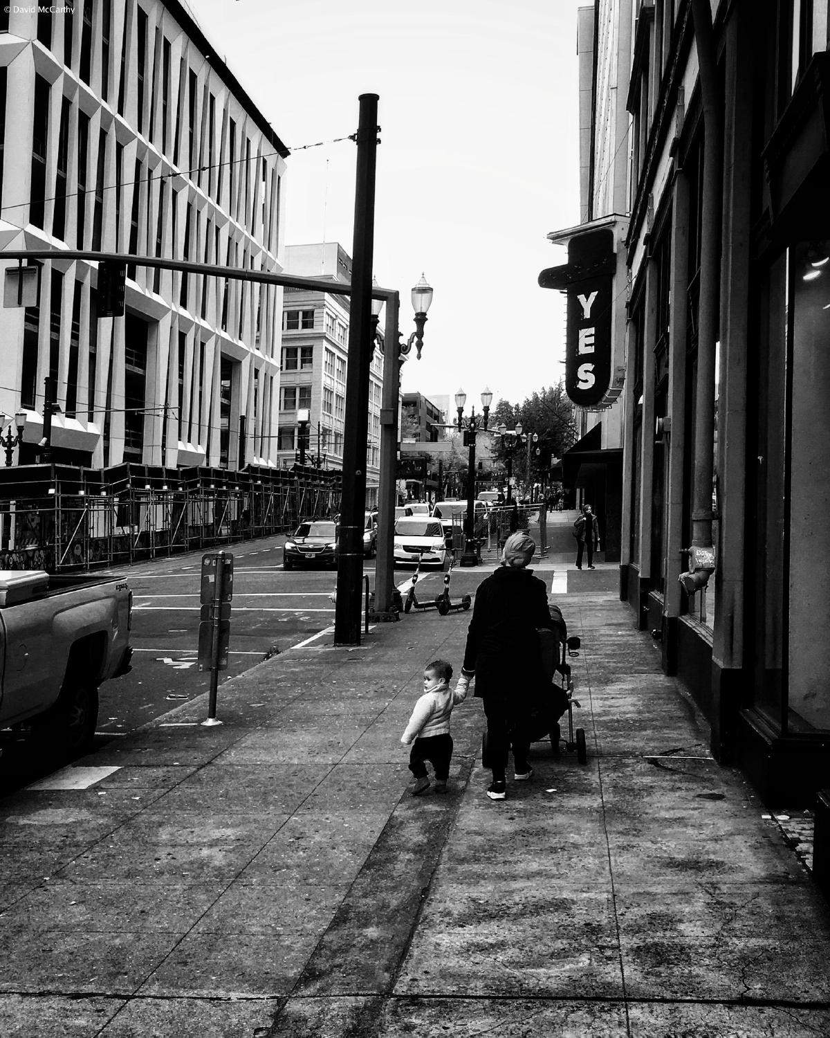 Black and white photograph of a parent walking on a sidewalk while holding a young child's hand and pushing a stoller.