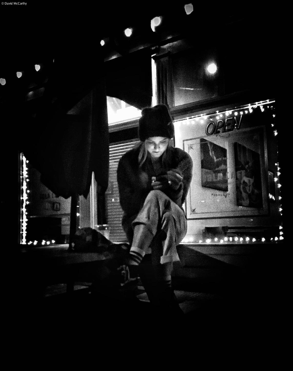 Black and white photograph of a woman sitting on a table outside, smoking and looking at the screen of a device in her hand.