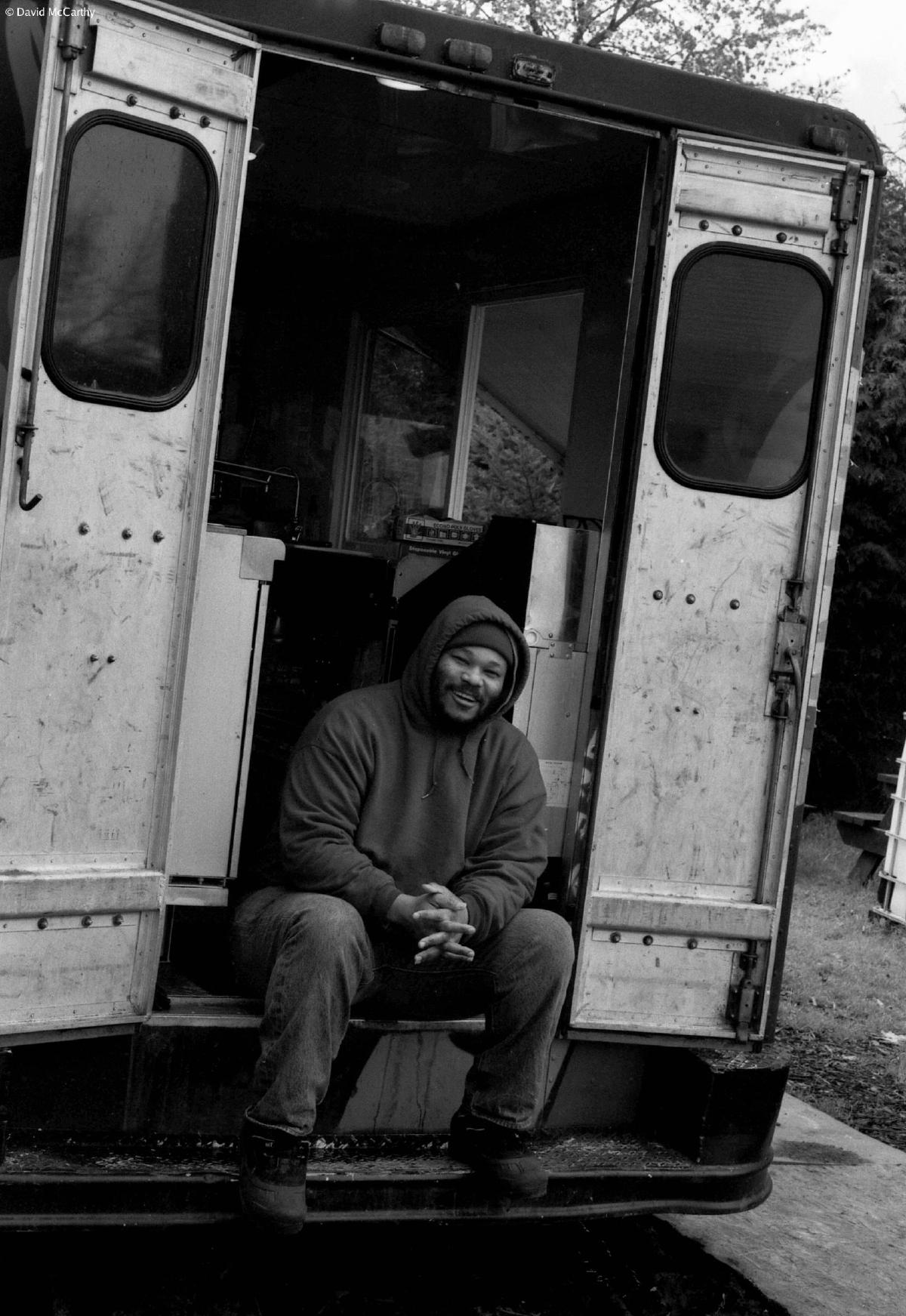 Black and white photograph of a smiling man in a hooded sweatshirt sitting in the back of a panel truck.