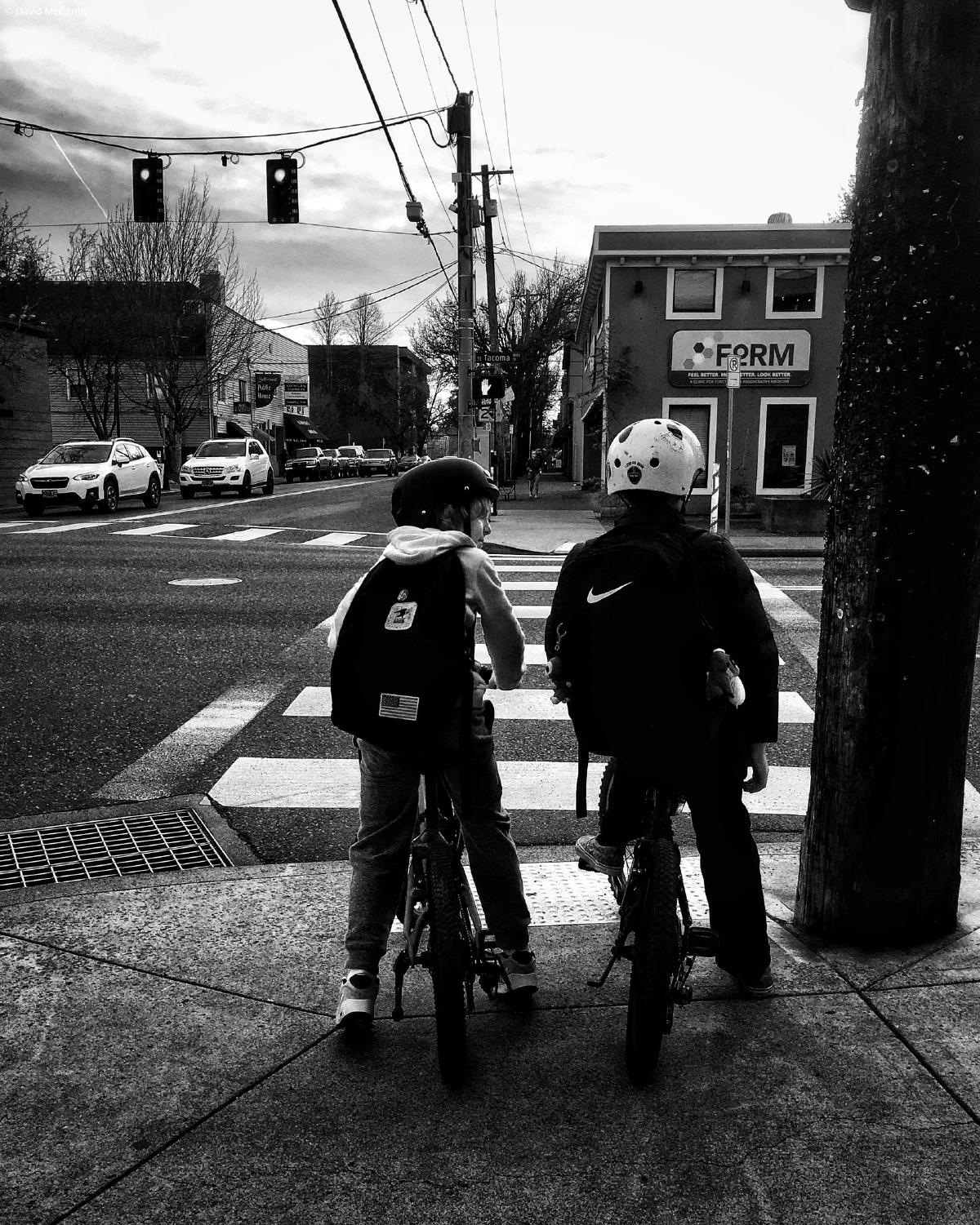 Black and white photograph of two kids on bicycles with helmets and backpacks, waiting to cross SE Tacoma Street at SE 13th Avenue in Portland, Oregon.