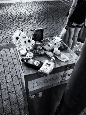 Black and white photograph of a newspaper distribution box in downtown Portland for The Epoch Times. The box is covered with empty packs of cigarettes, an empty pint of vodka, an bottle of liquid iron supplement, and flowers.