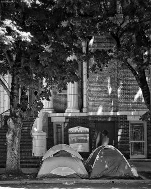 Black and white photograph of two dome camping tents on a sidewalk outside of First Christian Church in downtown Portland Oregon. A sign in the church display behind the tents reads 'Be Loved'.