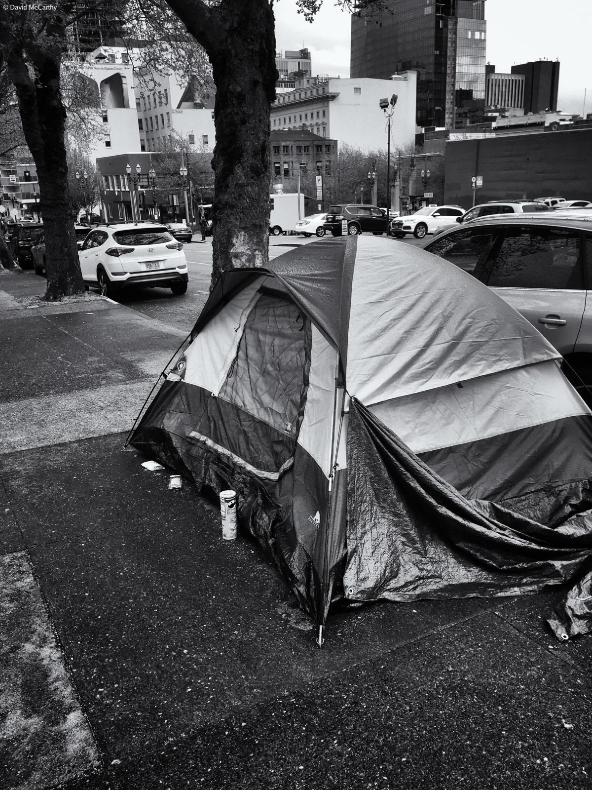 Black and white photograph of a visibly wet camping tent on a sidewalk in Portland Oregon. A can of Pringles brand potato chips sits near the entrance of the tent.