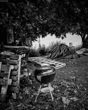 Black and white photograph of a makeshift campsite near the Willamette River in Portland Oregon. Many wooden pallets are visible, along with a charcoal grill, a shopping cart filled with discarded items, and a dome camping tent near the river.